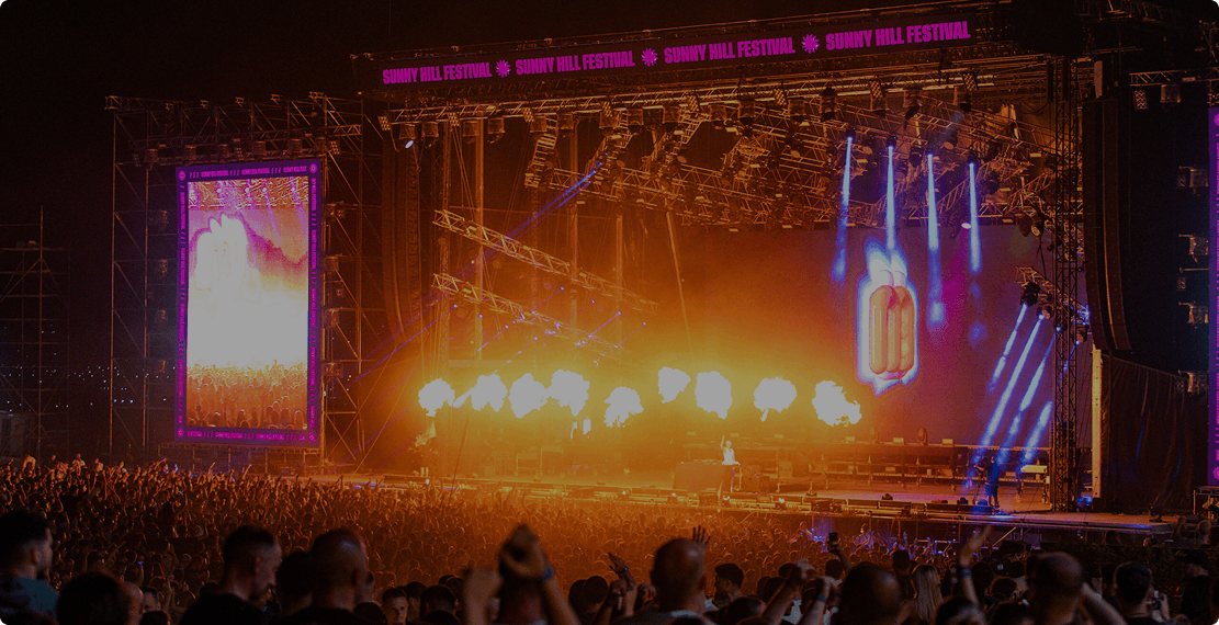 Large outdoor concert stage with bright lights, erupting flames, and a cheering crowd at night.